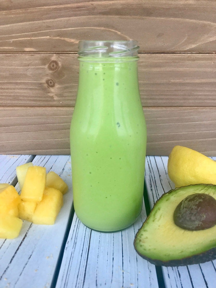 An avocado smoothie in a glass bottle next to pineapple and half an avocado.