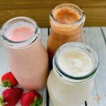 Three glass bottles with milkshakes. One chocolate, one strawberry, and one vanilla, with 3 strawberries next to them.