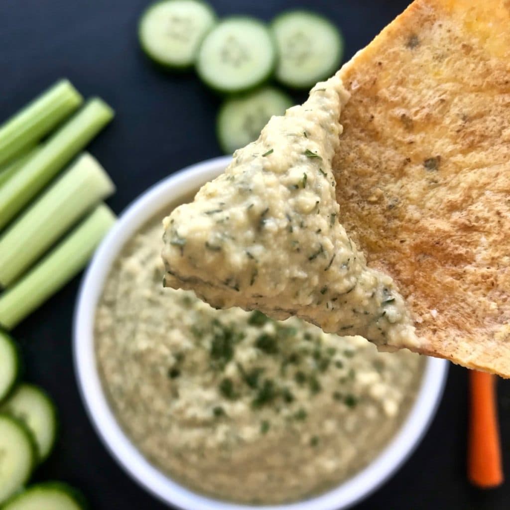 A tortilla chip with dill pickle hummus on it, with more hummus and veggies in the background.