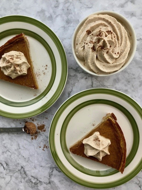 A small bowl of pumpkin whipped cream and two slices of pumpkin pie on separate plates with pumpkin whipped cream on top.