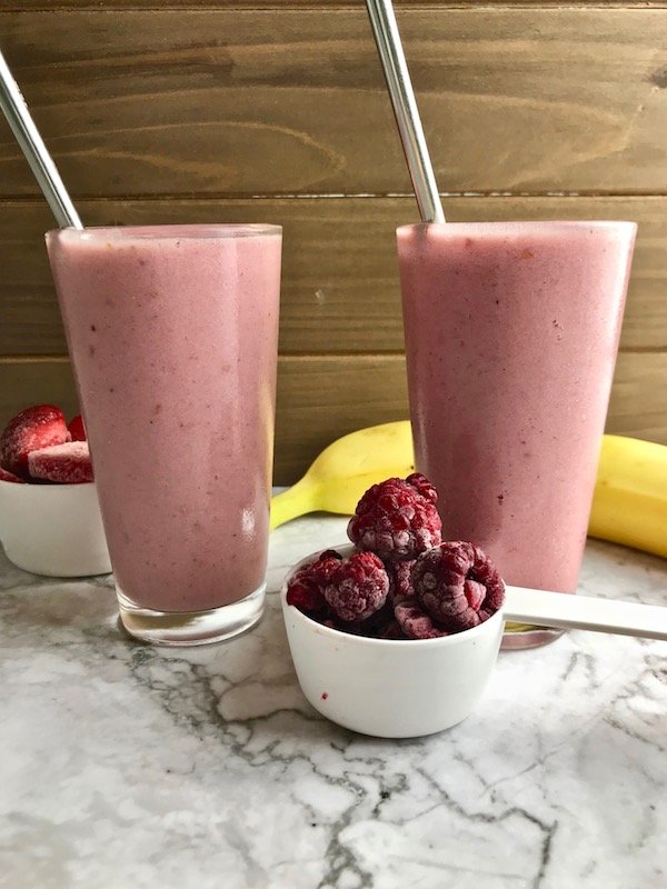 Two strawberry banana raspberry smoothies with ingredients on the table. 