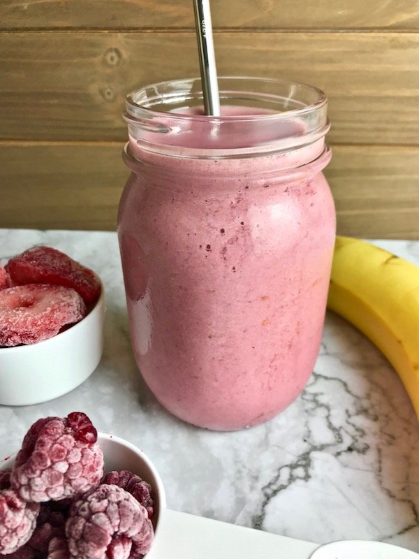 Strawberry banana raspberry smoothie in a clear mason jar with ingredients on the table. 