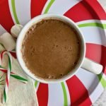 Cup of cinnamon hot chocolate on a red, white, and green striped plate, next to marshmallows and candy canes.