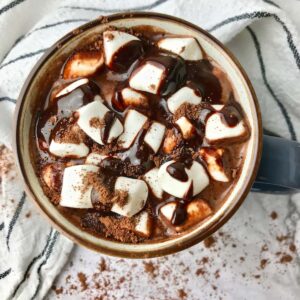 A mug of hot chocolate topped with mini marshmallows and chocolate sauce.