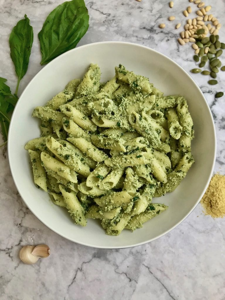 Pesto sauce on penne pasta in a white bowl, with pesto ingredients in the background. 