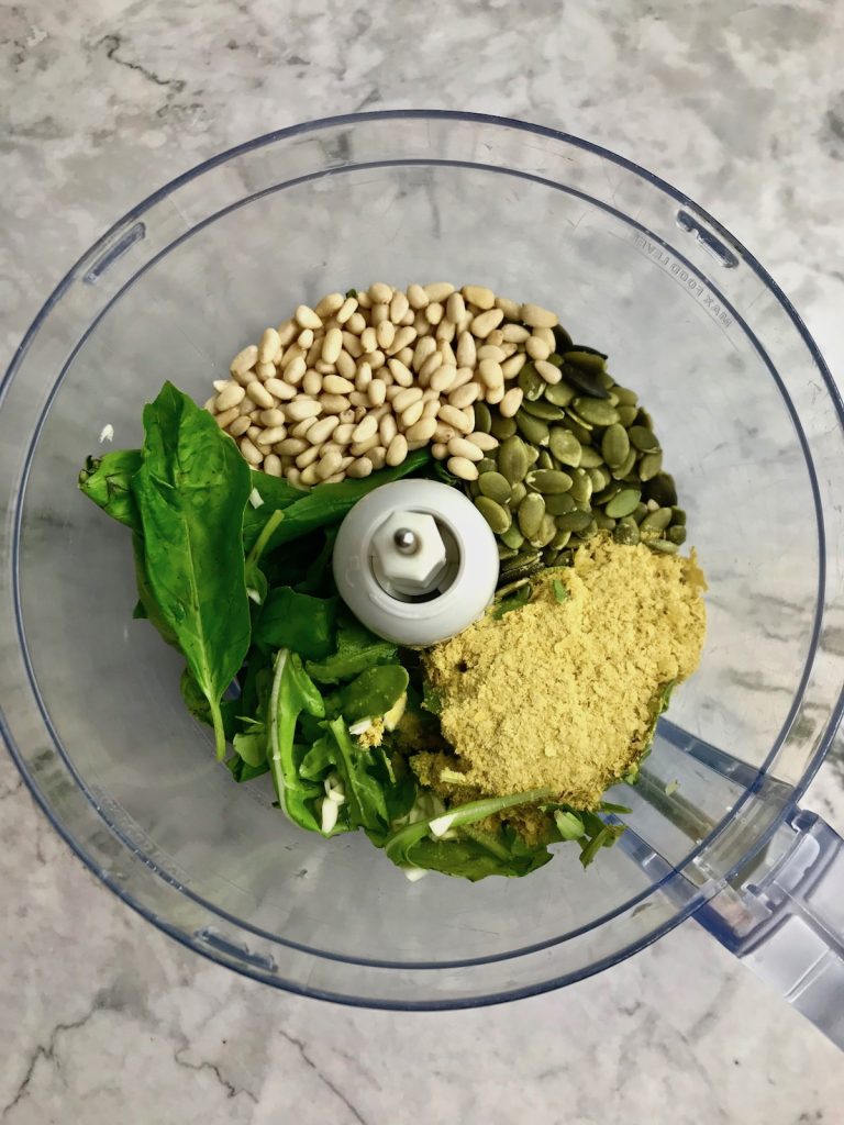 Ingredients for pesto in a food processor, including basil, arugula, pine nuts, pepitas, and nutritional yeast. 