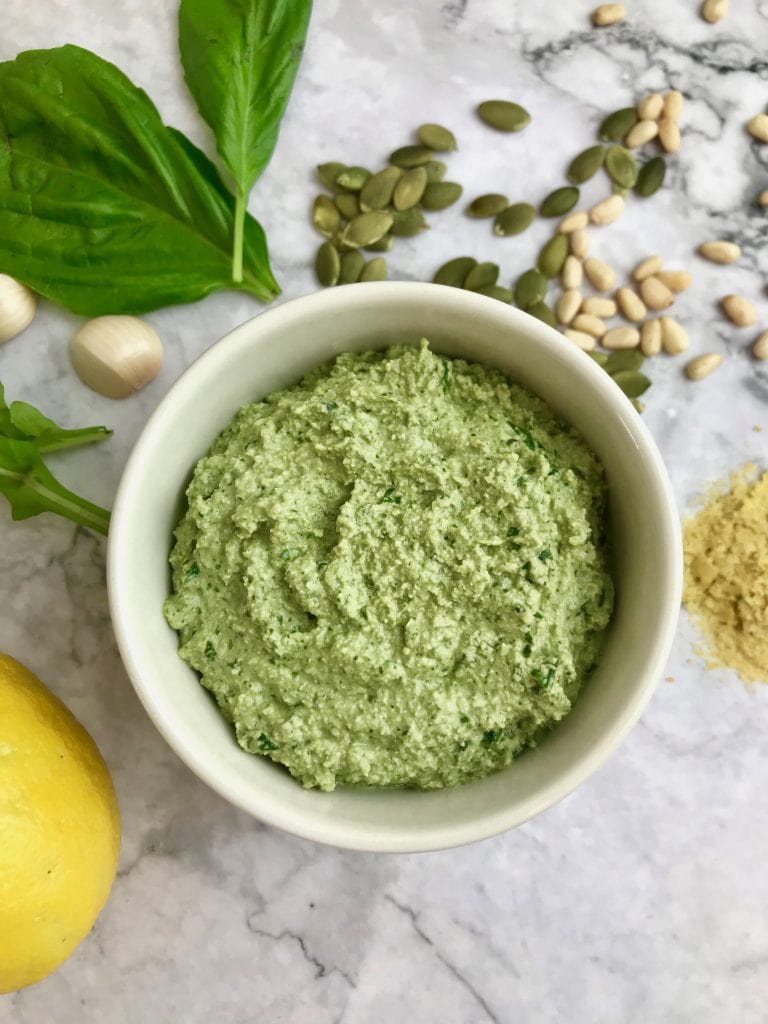 Oil-free vegan pesto in a white dish with ingredients on the table surrounding it, including basil, arugula, pepitas, pine nuts, nutritional yeast, and a lemon.