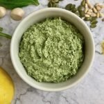 vegan pesto in a small white dish, surrounded by pine nuts, pepitas, garlic, basil, and lemon on the table.