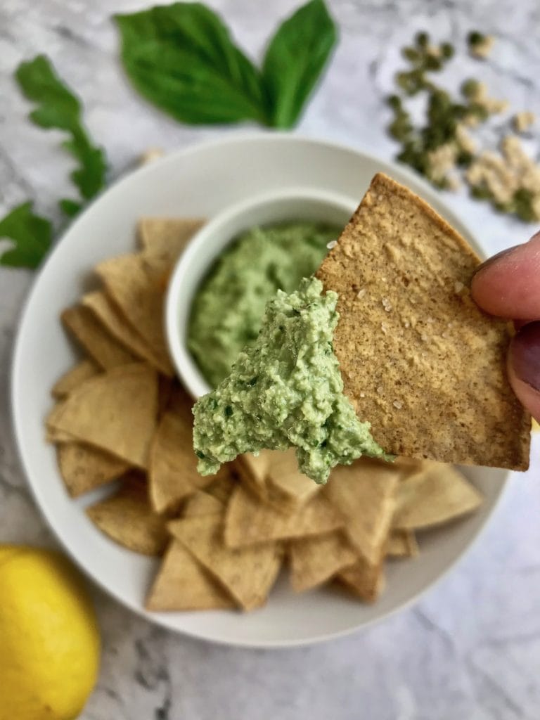 Close up of pesto on a tortilla chip, with ingredients in the background. 