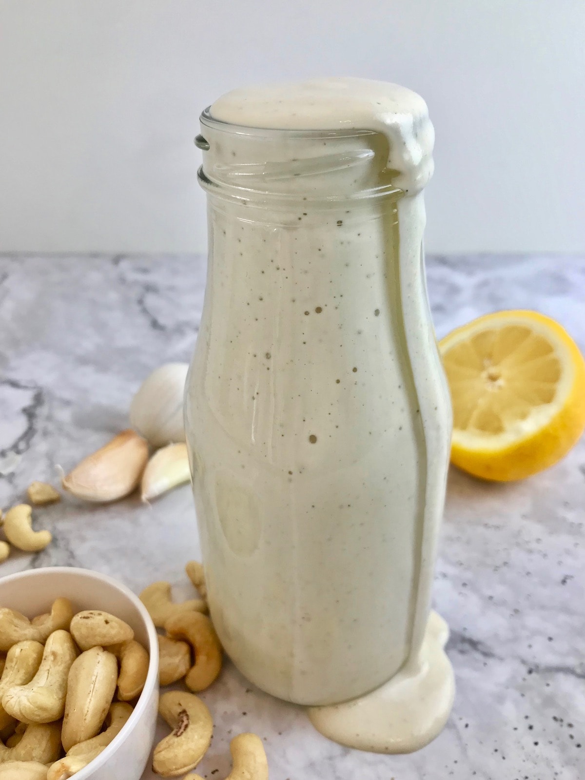 Bottle of white caesar dressing overflowing on a table with cashews, garlic cloves, and half a lemon.