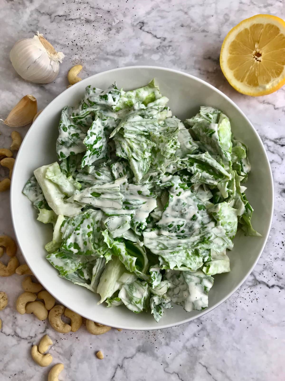 A bowl of romaine lettuce topped with white creamy dressing, and cashews, garlic, and lemon on the table. 