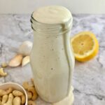 Bottle of white caesar dressing filled to the top with cashews, garlic cloves, and half a lemon on the table.