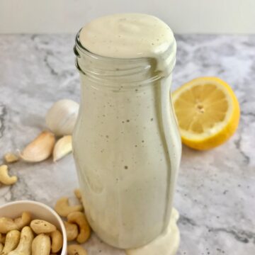 Bottle of white caesar dressing filled to the top with cashews, garlic cloves, and half a lemon on the table.