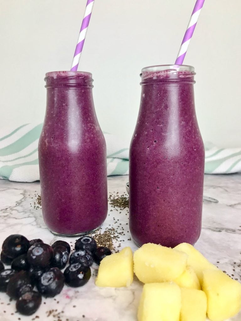 two purple smoothies in glass milk jars with blueberries and pineapple chunks in front of them. 