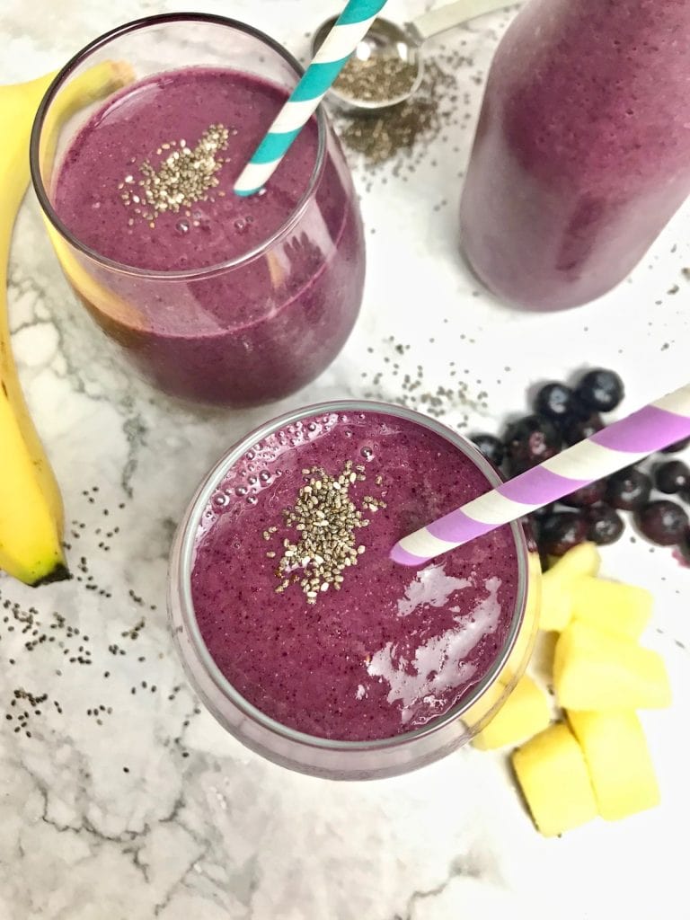 Overhead view of two purple smoothies with banana, pineapple, and blueberries on the table.