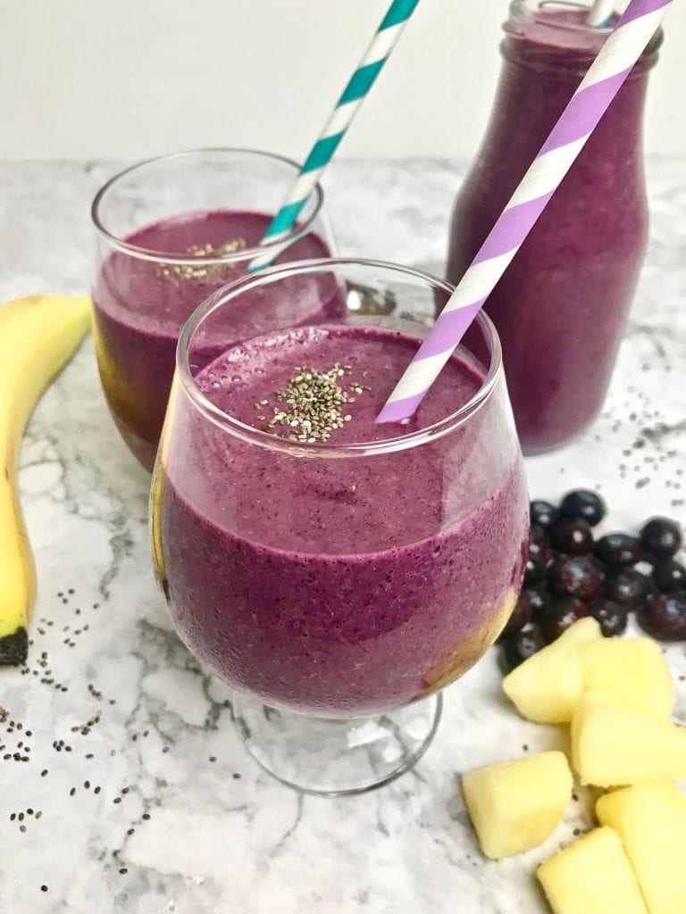 Purple smoothie with a white and purple striped straw, on a table with a banana, pineapple chunks, and blueberries, and two more purple smoothies in the background.