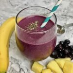 Purple smoothie in a stemless wineglass on a table with a banana, pineapple chunks, and blueberries.