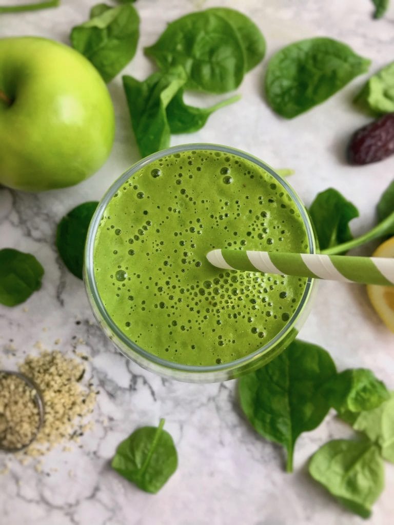 Overhead view of green smoothie with a green and white striped straw, surrounded by smoothie ingredients on the table. 