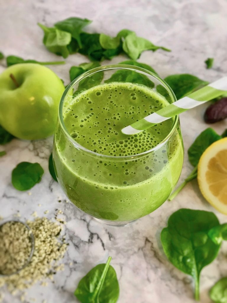 Green smoothie surrounded by ingredients such as spinach, apple, lemon, and hemp seeds. 