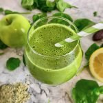 Green smoothie in a stemless wineglass, surrounded by spinach, green apple, lemon, and hemp seeds.