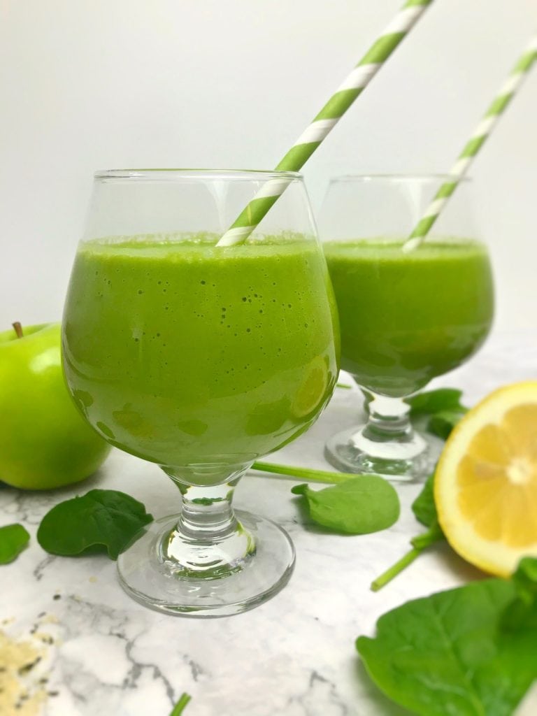Two green smoothies in short stemmed wine glasses with green and white striped straws and smoothie ingredients in the background. 