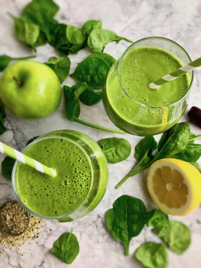 Overhead view of two green smoothies, with spinach, green apple, half a lemon, and hemp seeds surrounding them.