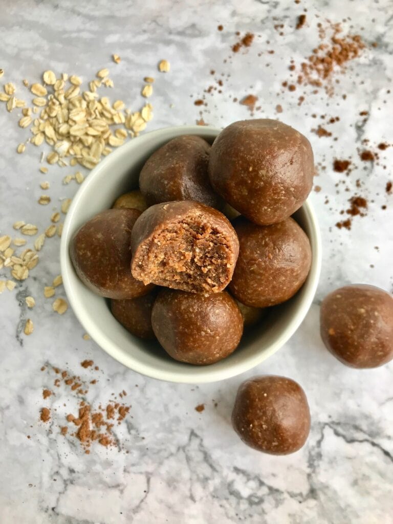 Brown chocolate cookie dough balls in a small white bowl, surrounded by oats and cocoa powder.