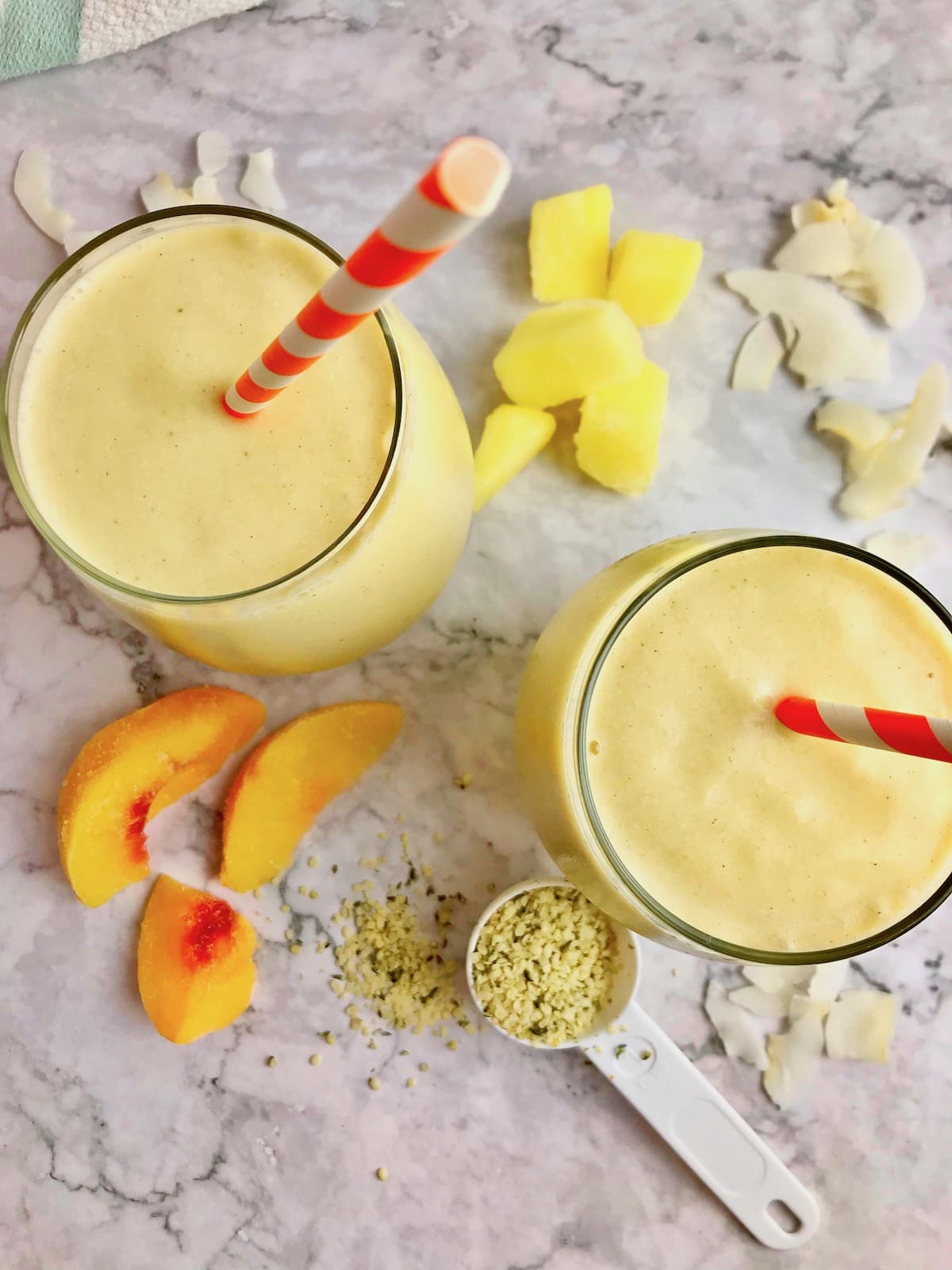 Two yellow pineapple peach smoothies with ingredients on the table, including fruit, coconut, and hemp seeds.