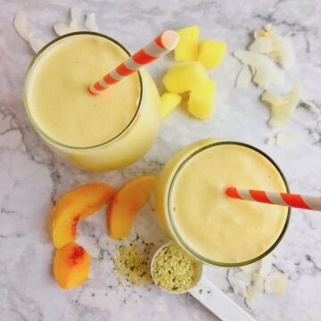 Two yellow smoothies with pineapple and peach slices on the table.