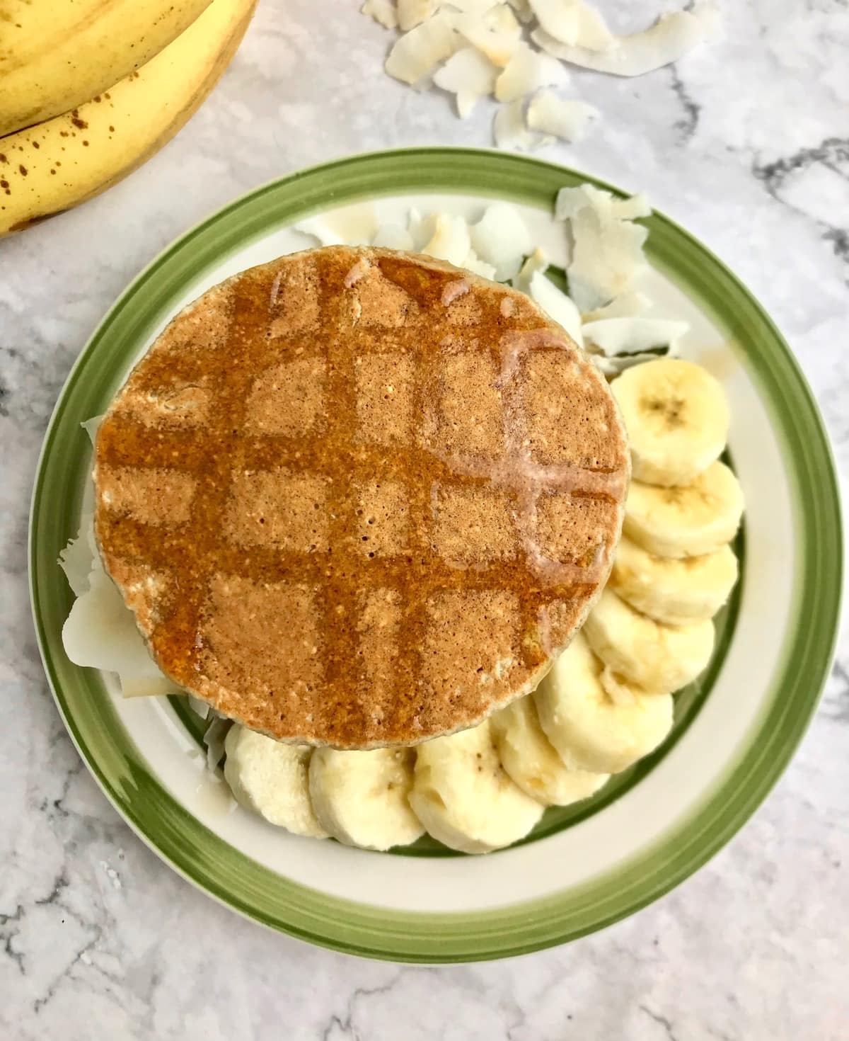 Overhead view of a pancake with maple syrup on top and banana slices  and coconut flakes next to it on a plate.