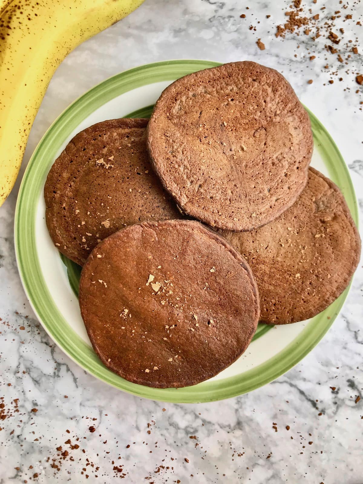 Four chocolate pancakes on a plate, with a banana on the side and cocoa powder sprinkled on top.