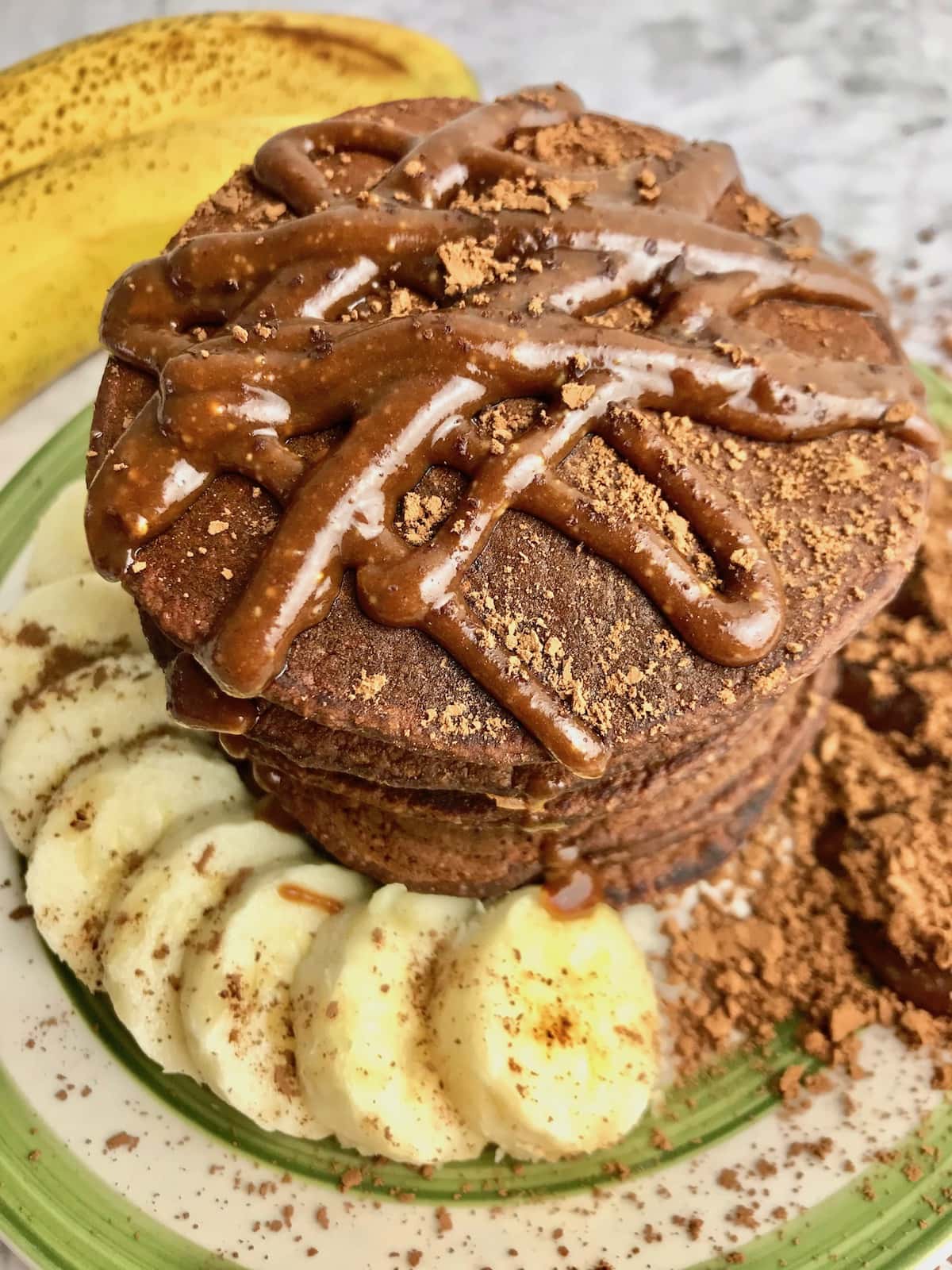 Stack of chocolate pancakes with chocolate syrup on top and banana slices on the plate.