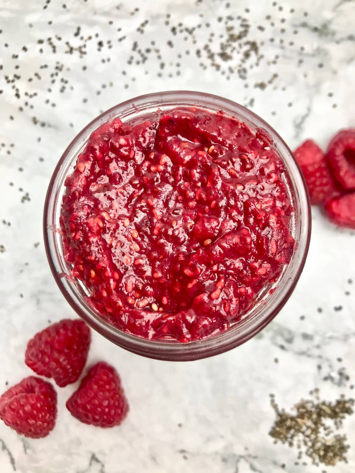 Overhead view of a container of red jam, with raspberries on the table in the background. 