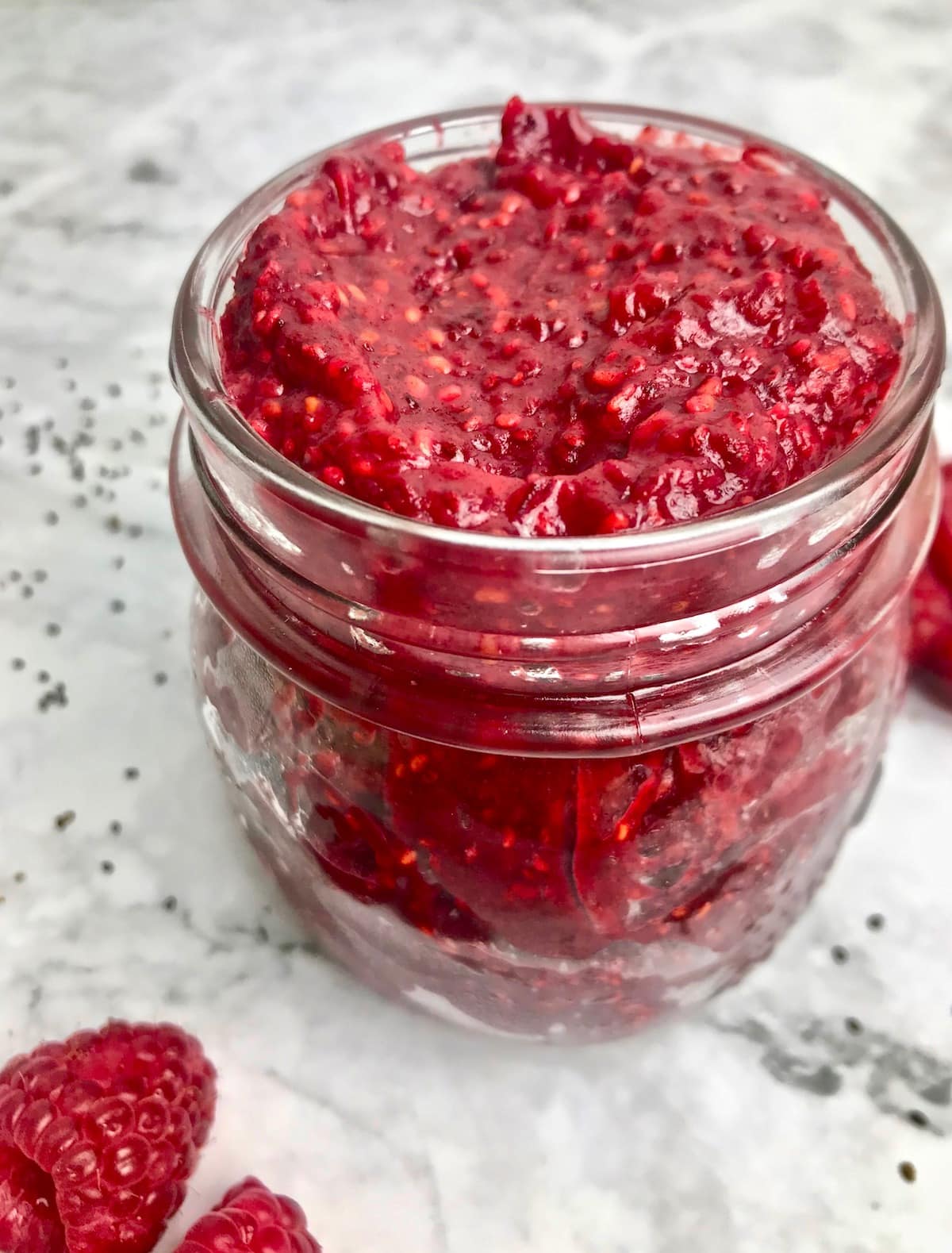 Small glass jar of red raspberry jam with raspberries on the table.