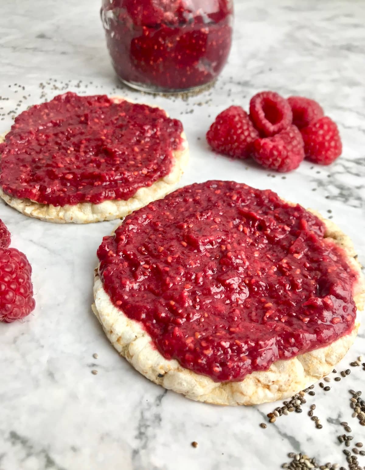 Rice cakes with red jam on top and raspberries on the table with a glass jar of jam in the background.