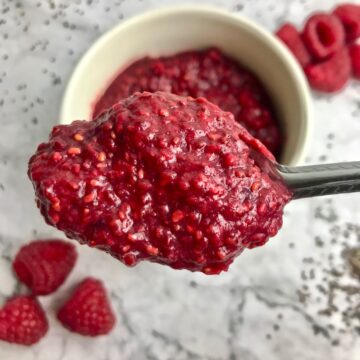 Close up of spoonful of raspberry jam with raspberries on the table in the background.