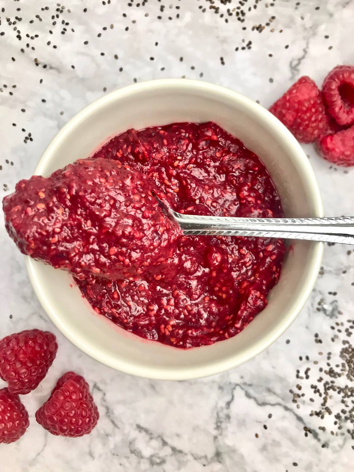 Spoonful of jam laying across a white dish of jam with raspberries on the table in the background.