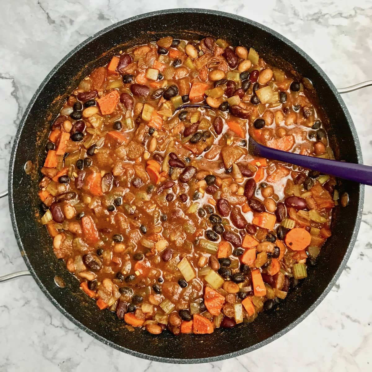 Vegan bean chili in a dutch oven pot with a purple serving spoon in it. 