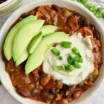 Bean chili in a bowl topped with vegan sour cream, avocado, and green onion.