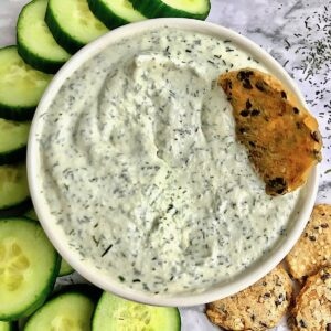 Tzatziki with a cracker dipped into it surrounded by more crackers and cucumber slices on a table.