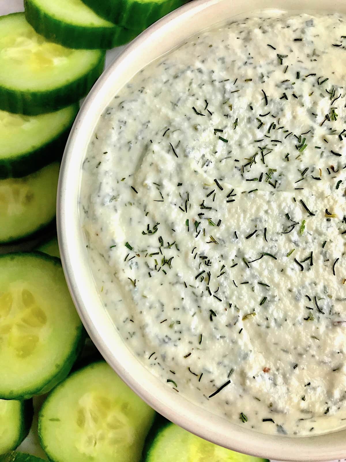 Half a bowl of vegan tzatziki dip with cucumber slices next to it.