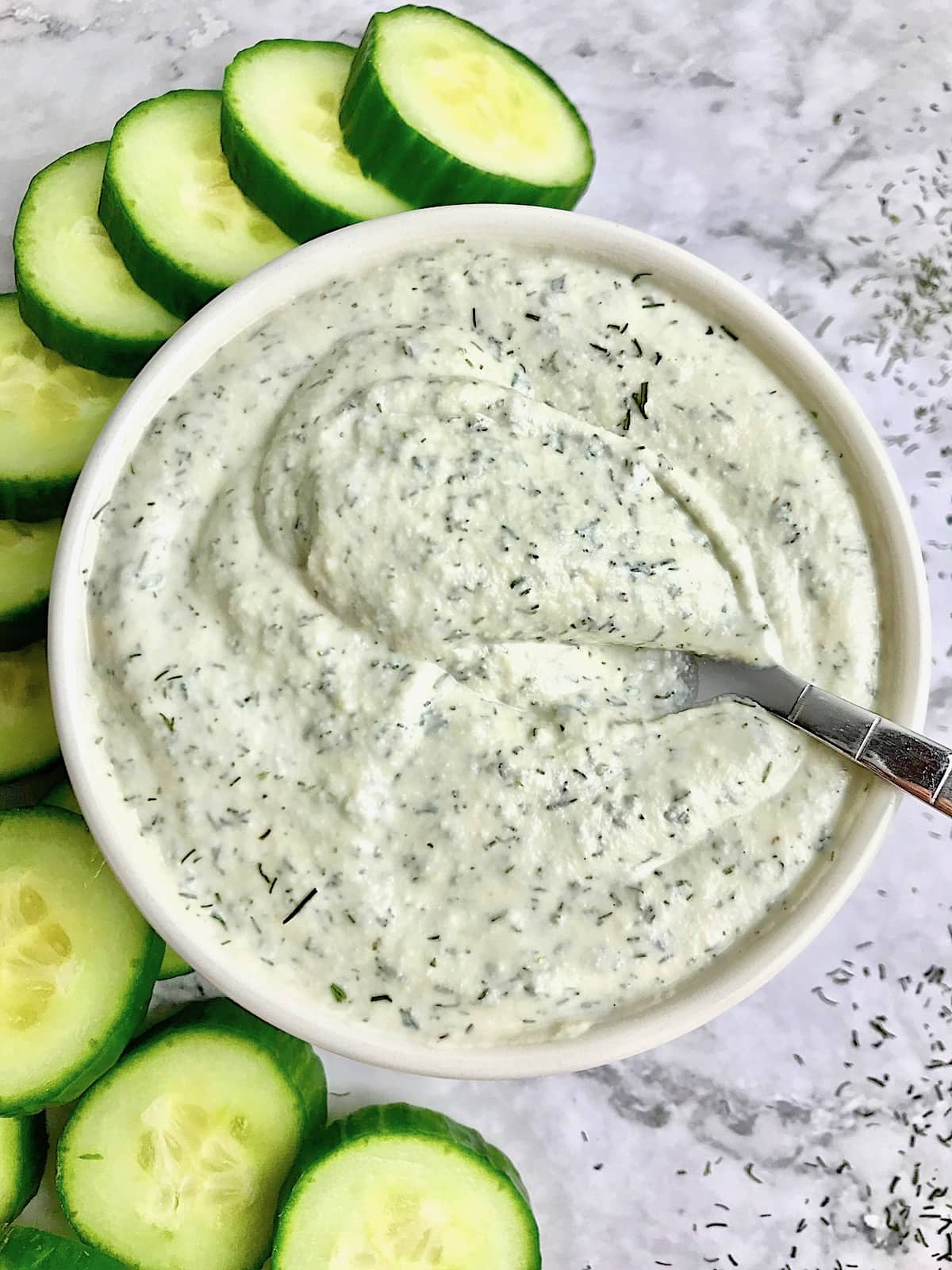 Tzatziki sauce in a white bowl with a spoon in it, sitting next to cucumber slices. 