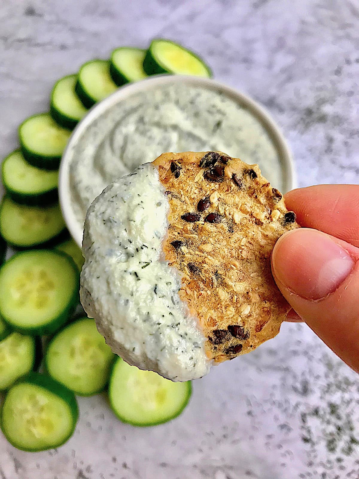 Close up of a cracker with tzatziki on it with a bowl of tzatziki and cucumber slices on a table in the background.