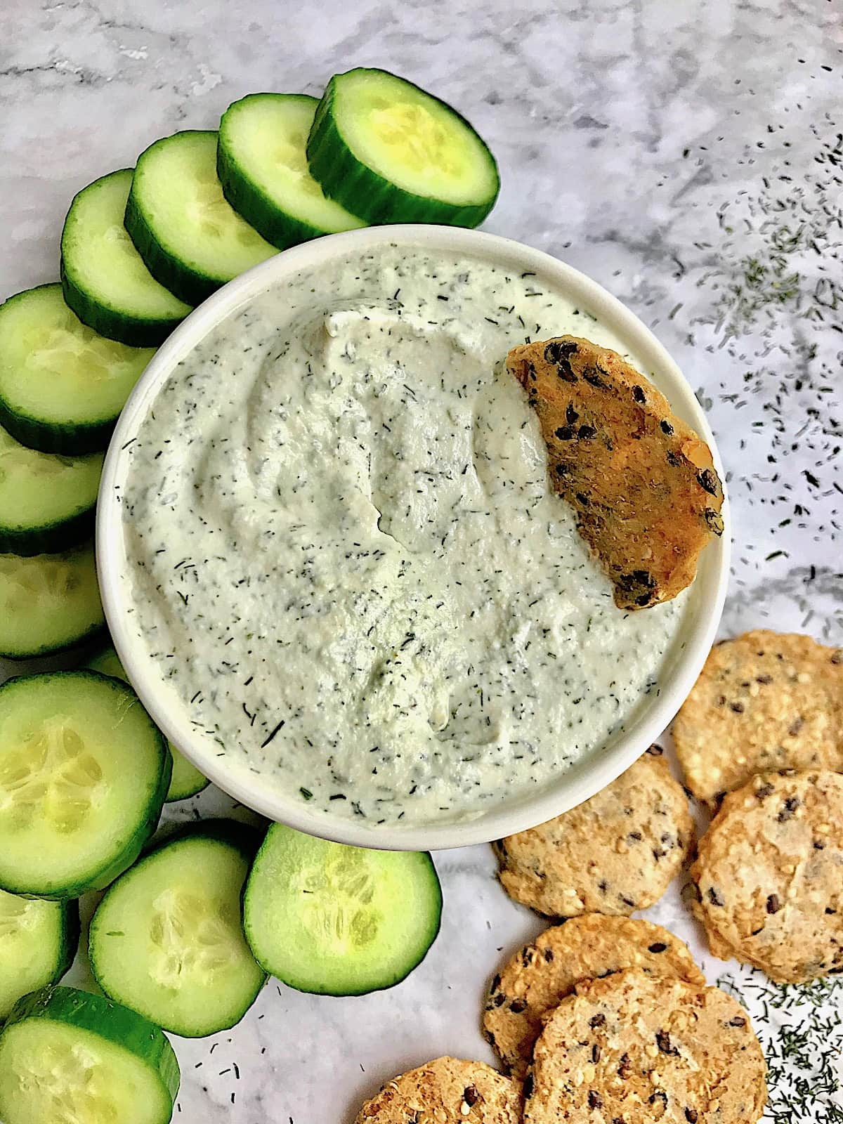 Tzatziki surrounded by cucumber slices and crackers on a table with dill sprinkled on it.