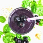 Overhead of a smoothie with blueberries on top and spinach leaves on the table in the background.