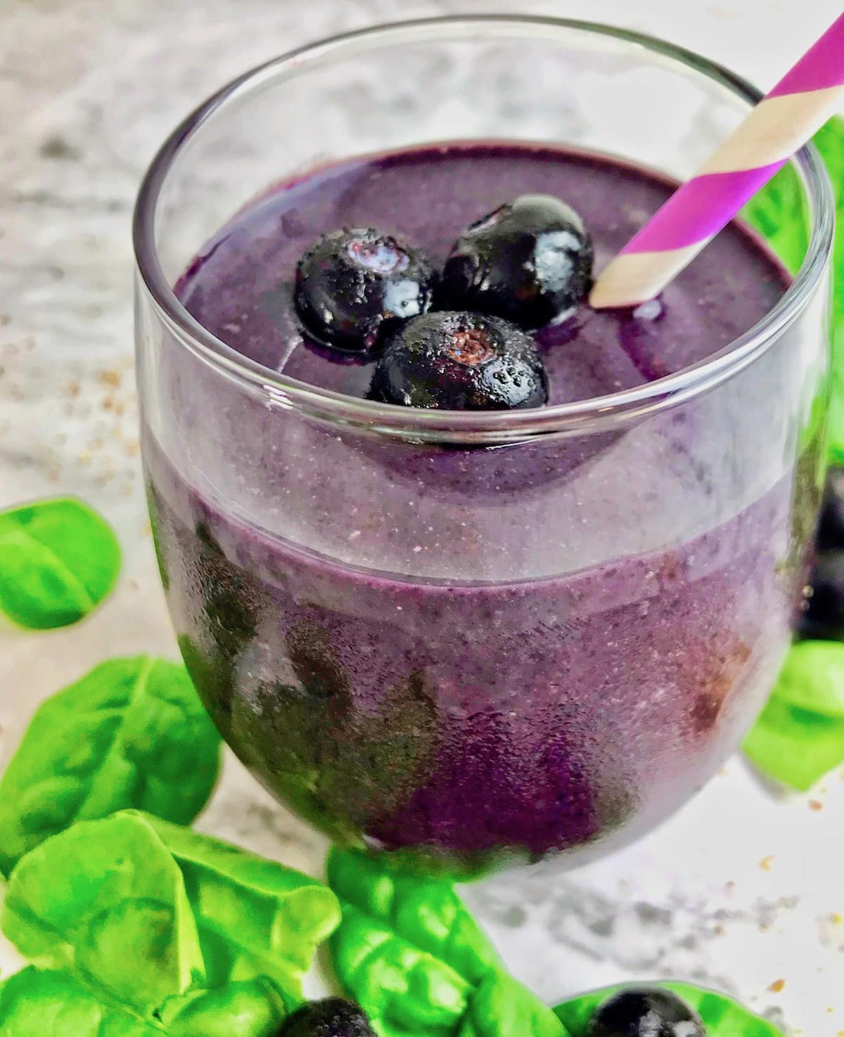 Blueberry smoothie in a stemless wine glass with blueberries on top and spinach surrounding the glass.