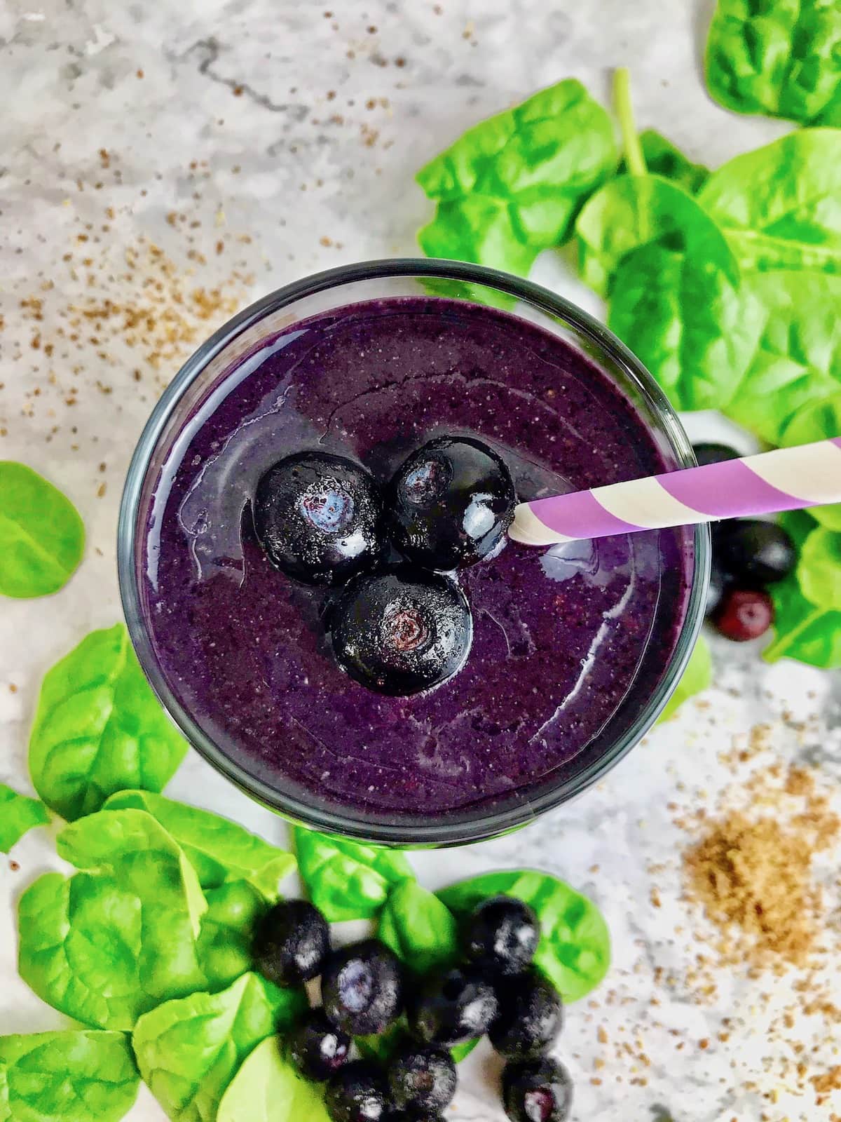 Overhead view of a purple smoothie with three blueberries on top, with spinach on the table in the background.