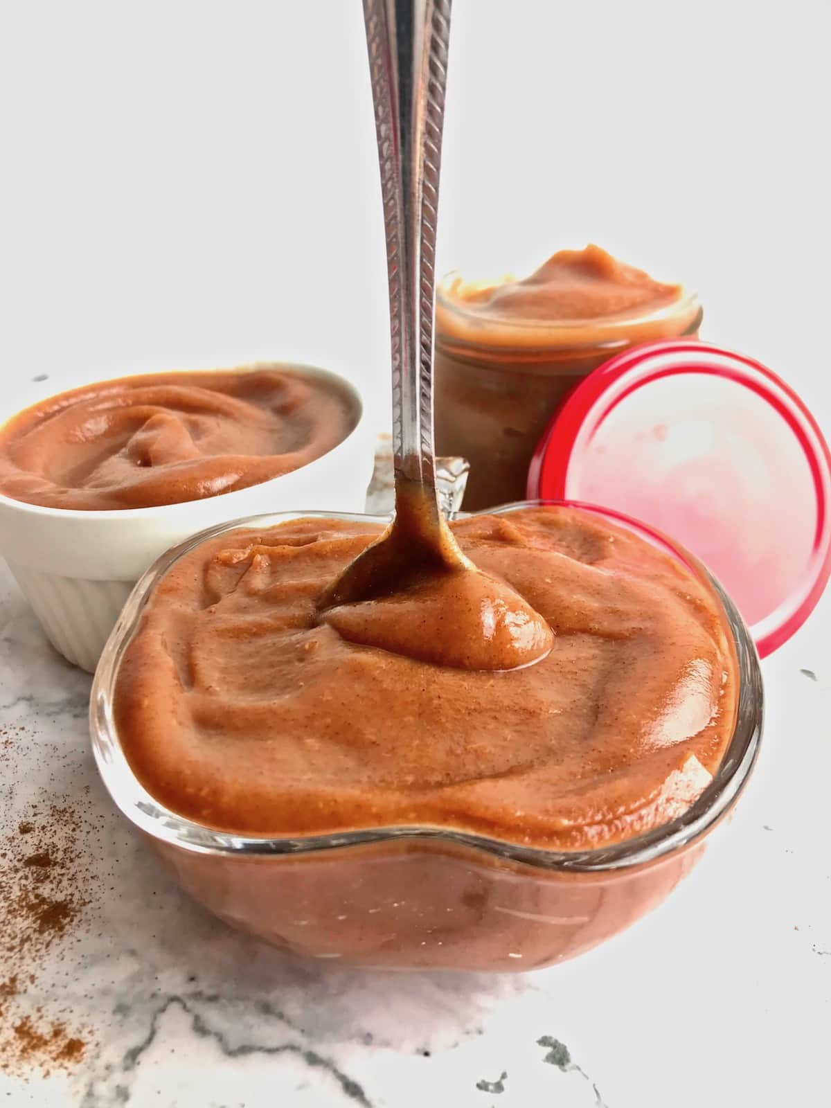 A jar of apple butter with a spoon standing up in it, with two more jars of apple butter in the background.