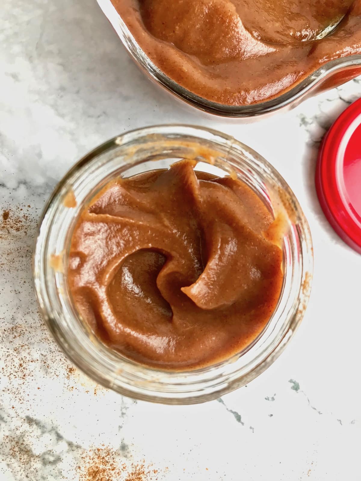 Apple butter in a round glass dish next to cinnamon and part of a red lid.