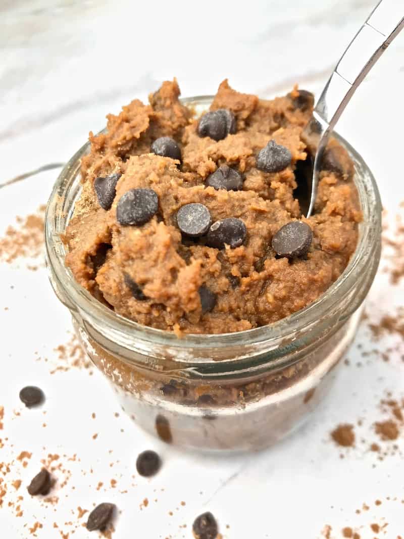 Chocolate chickpea cookie dough with a spoon in a glass jar with chocolate chips on the table.